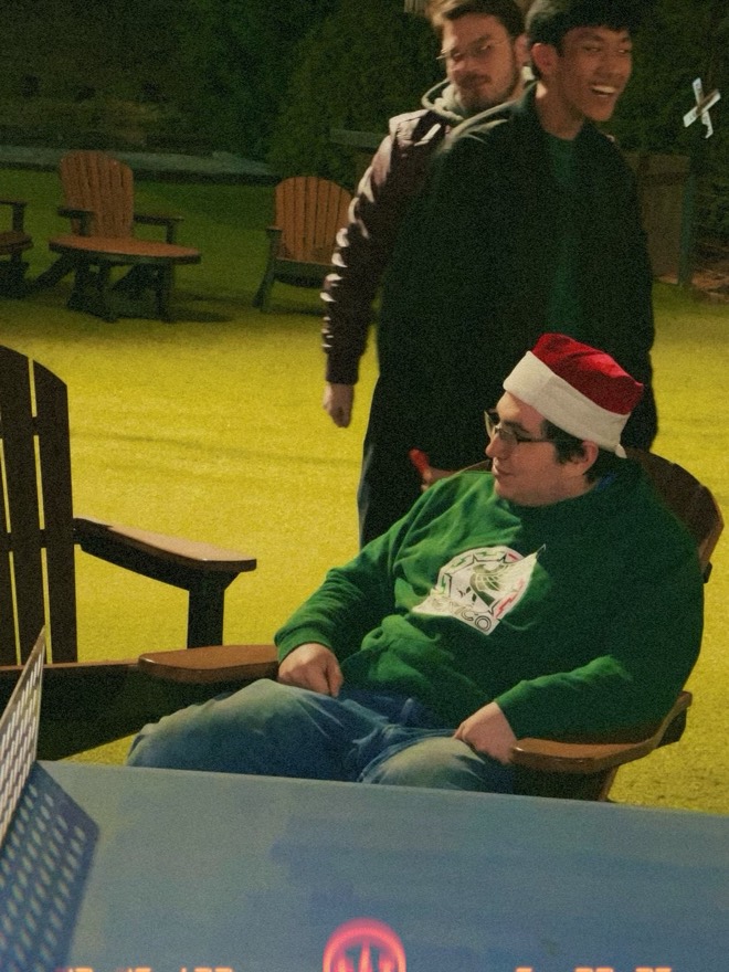Adam in a Santa hat sitting outdoors at night with friends behind him