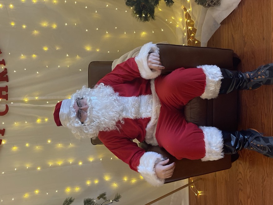 Adam in a full Santa Claus costume seated in an armchair in front of Christmas lights
